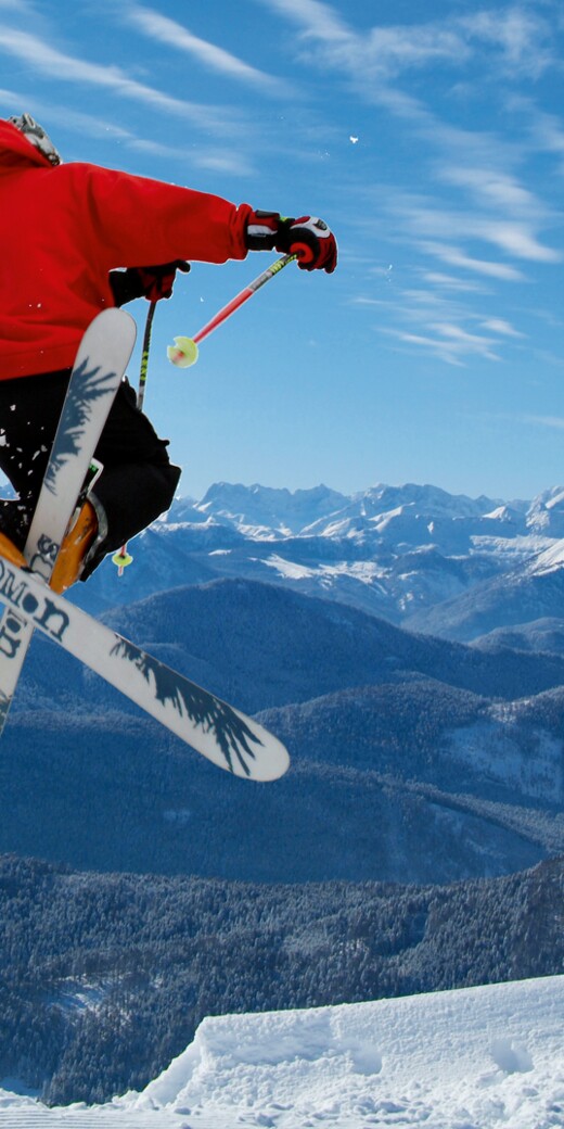 Ein Ski-Cross Fahrer springt gekreutzten Skiern in die Luft. Hinter ihm erstreckt sich ein beeindruckendes Bergpanorama im Winter.